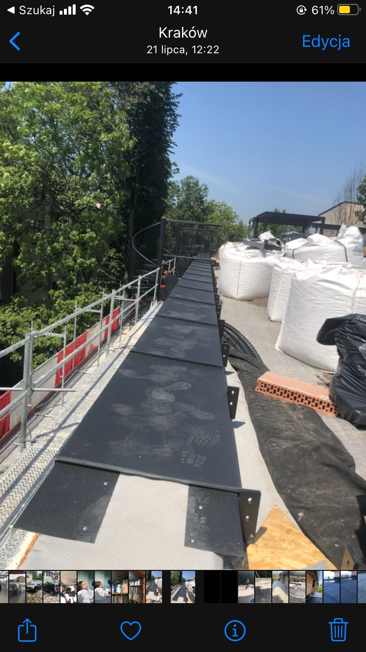 Czarna, tymczasowa ścieżka z płyt na dachu z widocznymi śladami butów, zabezpieczona barierkami, obok worki z materiałami budowlanymi i cegły.