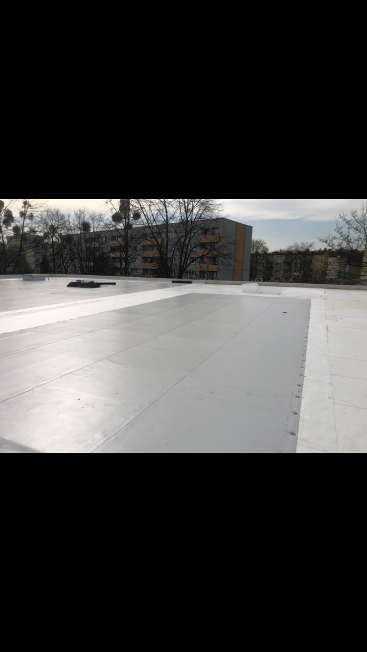 Szary, płaski dach z widocznymi łączeniami i elementami mocującymi, w tle budynek mieszkalny i drzewa bez liści.