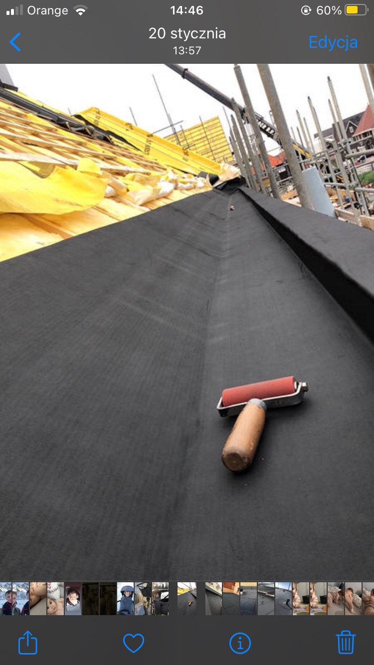 Czarna papa na dachu w trakcie montażu, widoczny wałek dociskowy z drewnianą rączką, w tle żółta membrana i rusztowania.