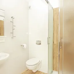 Jasna łazienka z umywalką, toaletą, prysznicem z przesuwanymi drzwiami i drewnianym panelem wewnątrz kabiny, oraz wieszakiem na ręczniki.