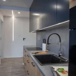 Nowoczesna kuchnia z szarymi szafkami, drewnianymi frontami dolnymi, czarnym zlewem i designerską lampą liniową na ścianie, widok na korytarz.