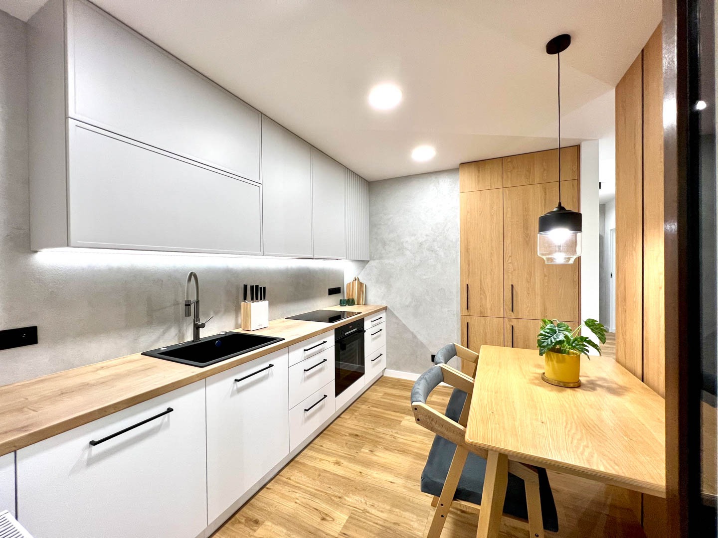 Nowoczesna kuchnia z białymi szafkami, drewnianym blatem i podłogą, szarymi ścianami, czarnym zlewem i armaturą, oraz drewnianym stołem jadalnym z rośliną w żółtej doniczce, oświetlona wiszącą lampą.