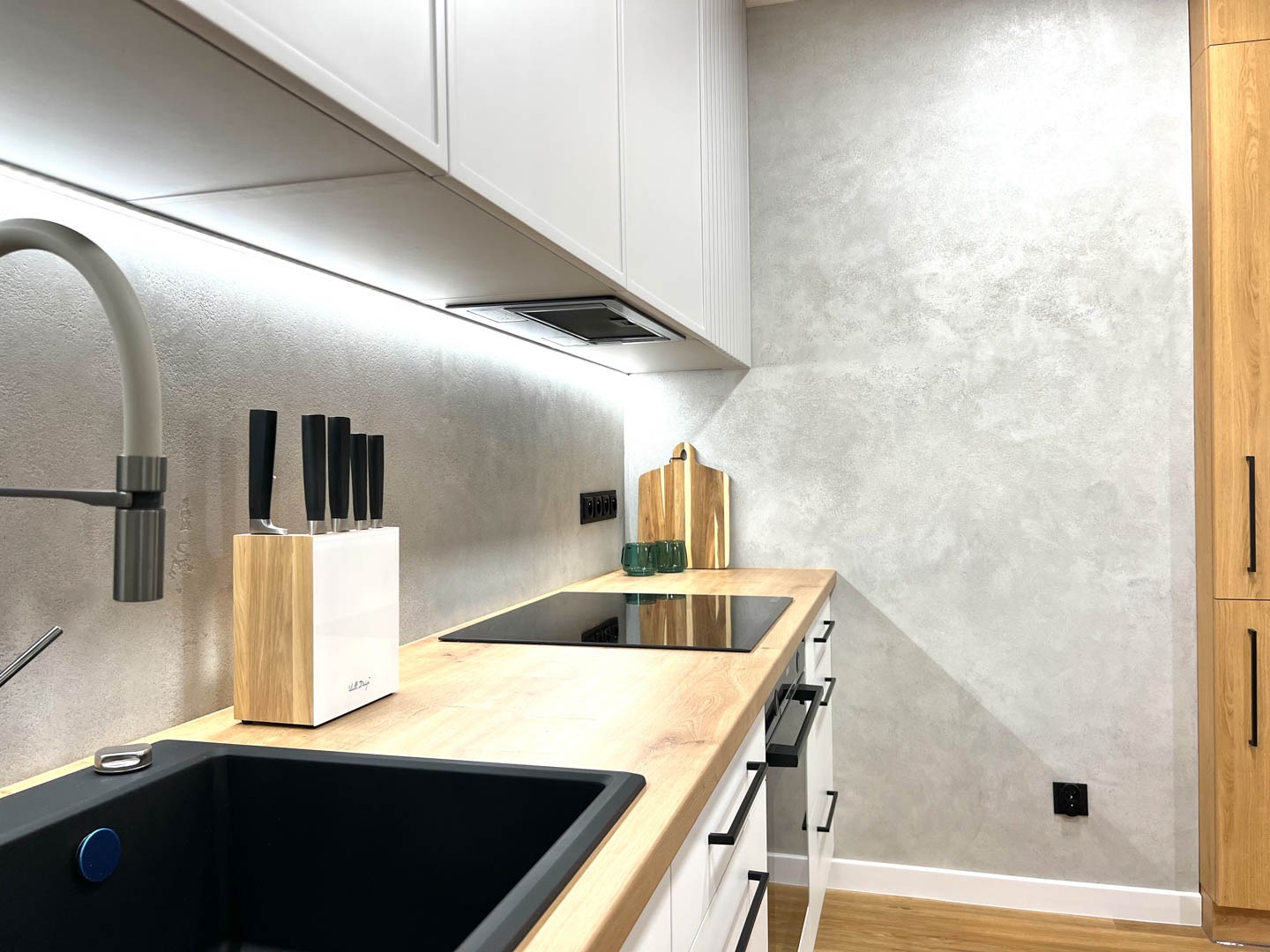 Nowoczesna kuchnia z szarą ścianą o fakturze betonu, drewnianym blatem, czarnym zlewem i białymi szafkami, oświetlona listwą LED pod szafkami górnymi.