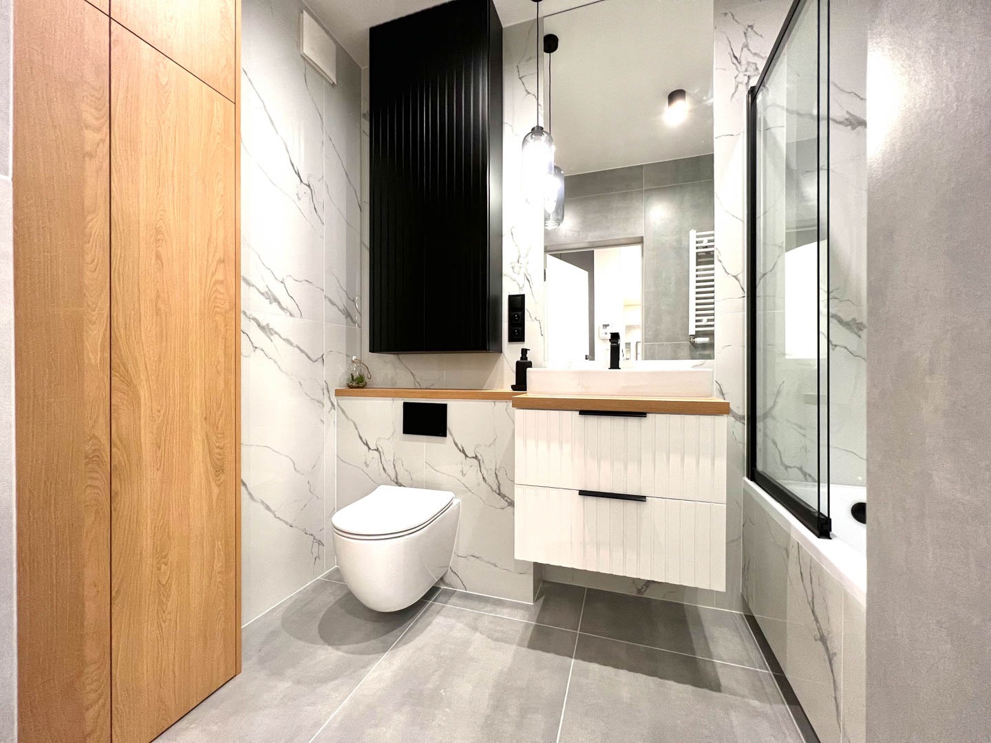 Nowoczesna łazienka z marmurowymi płytkami, wiszącą toaletą, szafką z umywalką na drewnianym blacie i czarnym grzejnikiem żeberkowym.