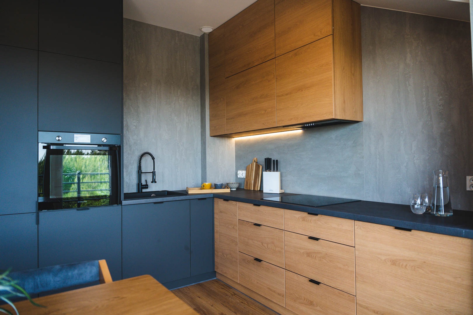 Nowoczesna kuchnia z szarymi ścianami, drewnianymi i grafitowymi szafkami, czarną armaturą i blatem. W tle widoczne okno z zielenią.