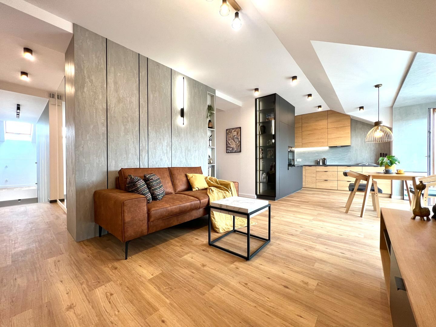 Nowoczesne wnętrze: salon z brązową skórzaną sofą, stolikiem kawowym z marmurowym blatem i widokiem na drewnianą kuchnię w tle, oświetlenie punktowe i podłoga z paneli.