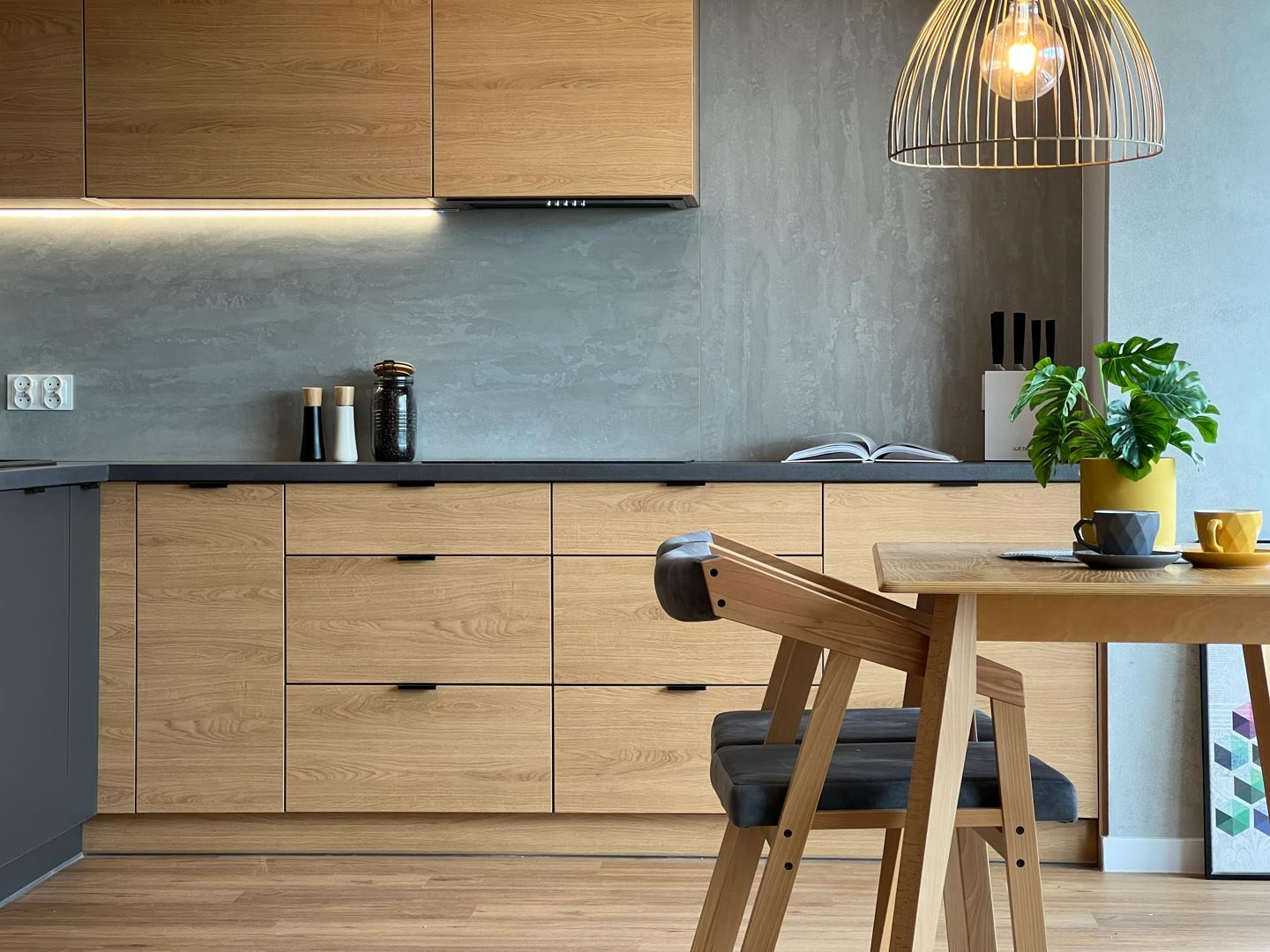 Nowoczesna kuchnia z drewnianymi frontami szafek, szarym blatem i ścianą o fakturze betonu, oświetlona lampą wiszącą z żarówką Edison, stół z drewnianymi krzesłami i dekoracjami.