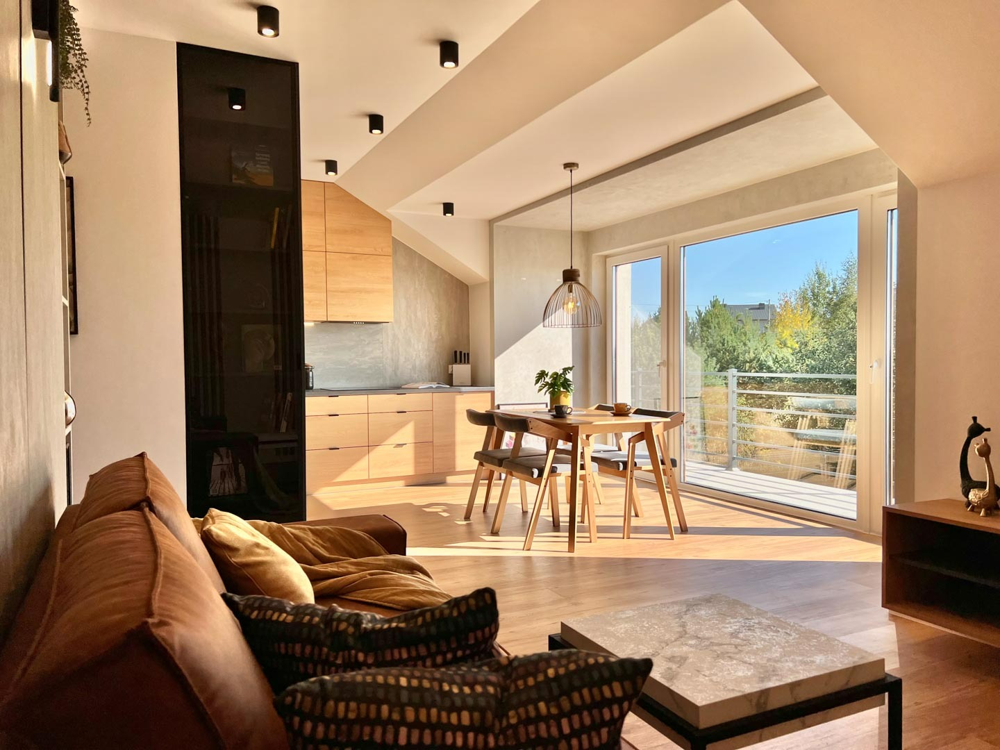 Jasne wnętrze mieszkania na poddaszu z drewnianą podłogą, widok na balkon i ogród, minimalistyczna kuchnia z drewnianymi szafkami i czarnym regałem, brązowa sofa z poduszkami i marmurowy stolik...