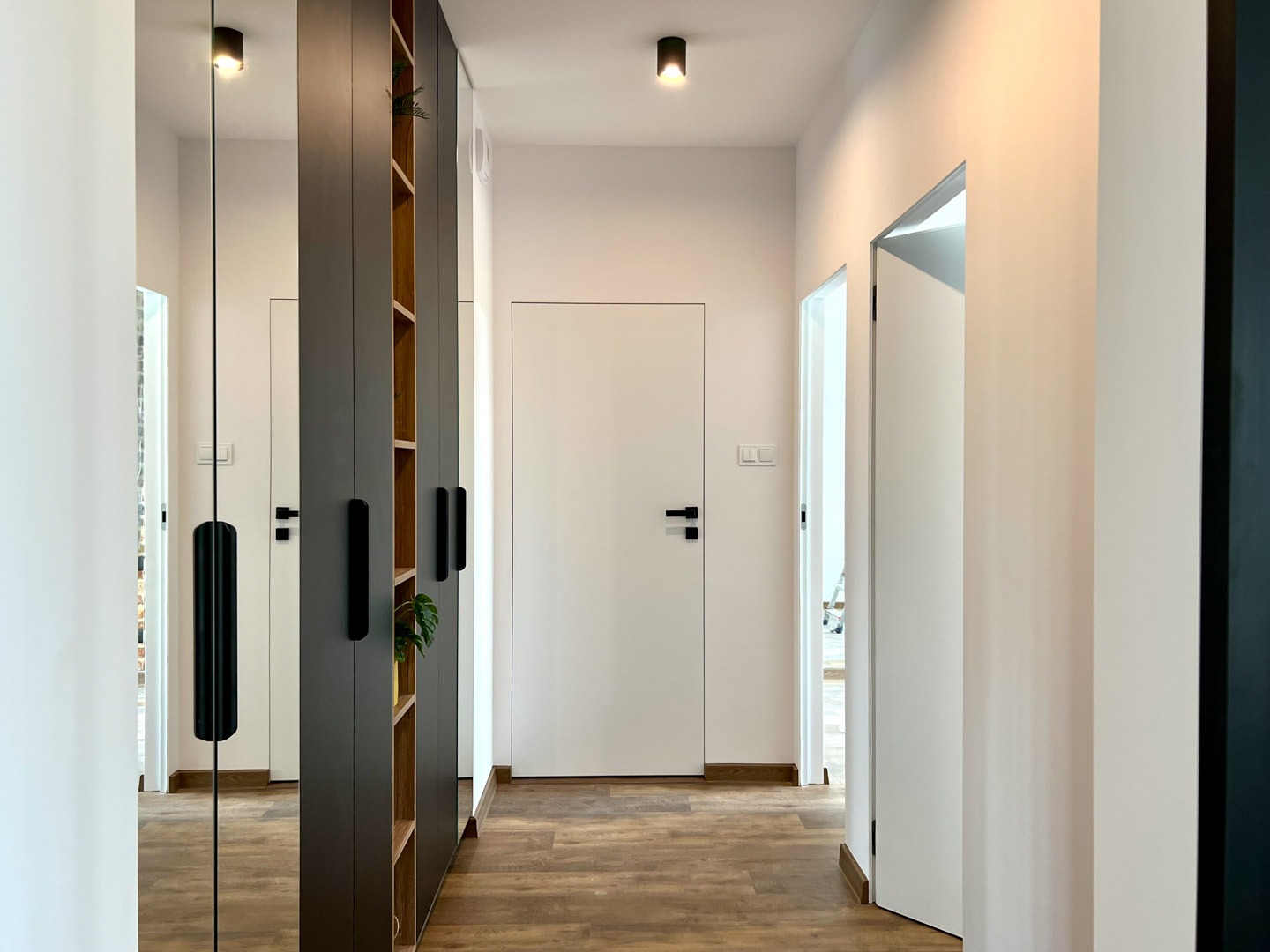 Nowoczesny korytarz z drewnianą podłogą, białymi ścianami i minimalistycznymi drzwiami, z szafą w kolorze antracytowym z otwartymi półkami i lustrem.