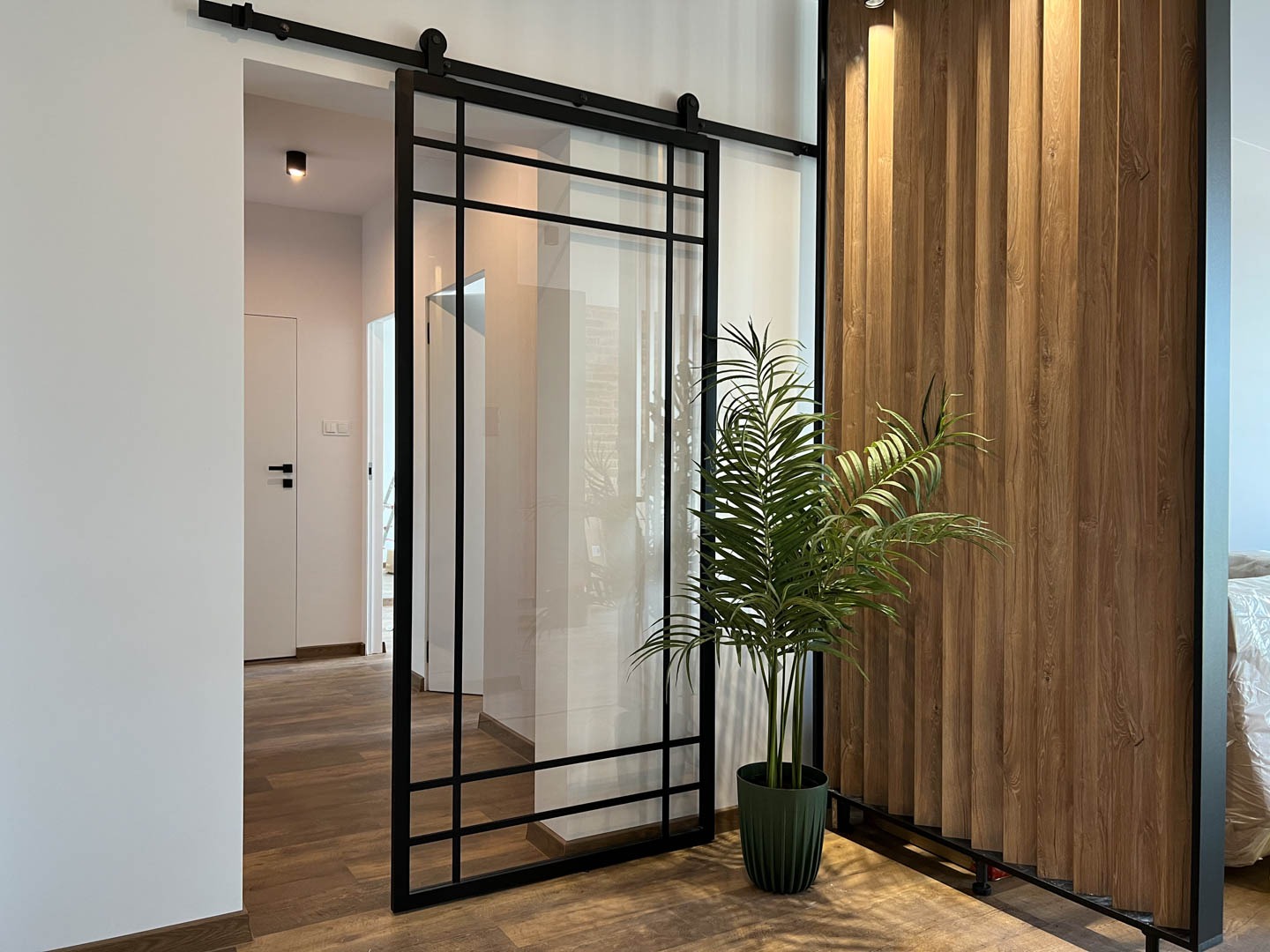 Nowoczesne, przesuwne drzwi loftowe z czarną ramą i szklanym wypełnieniem, zamontowane na szynie, widoczne z perspektywy wnętrza, z dekoracyjną ścianą z drewnianych paneli i rośliną doniczkową w tle.