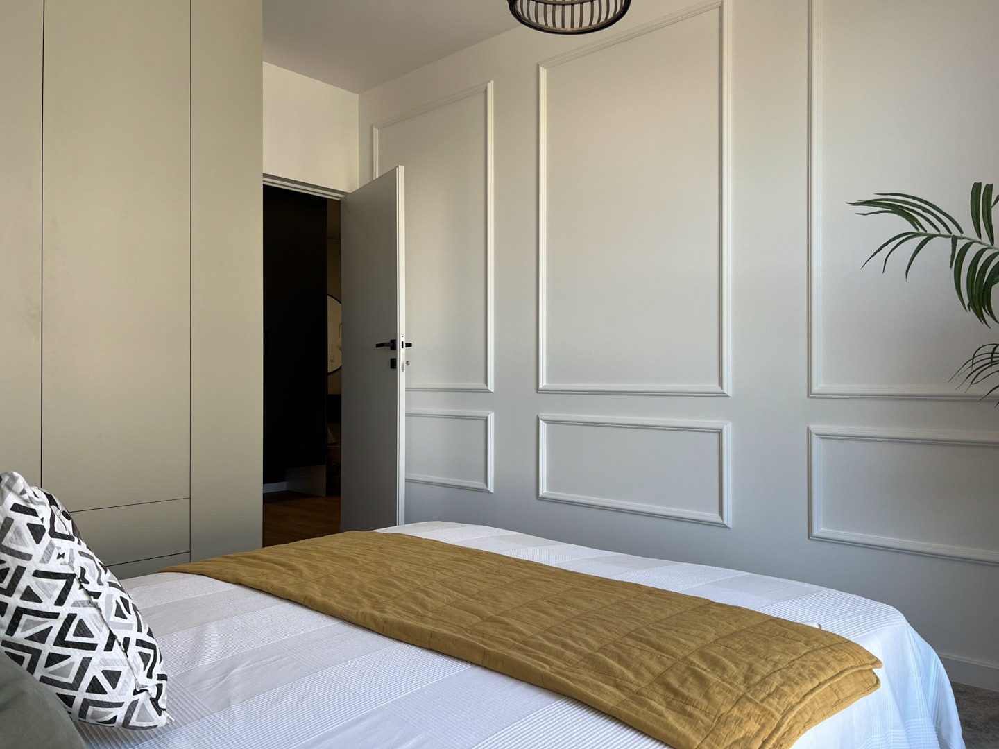 Jasna sypialnia z dekoracyjną sztukaterią na ścianach, otwartymi drzwiami i widocznym fragmentem garderoby oraz łóżkiem z geometryczną poduszką i musztardową narzutą.