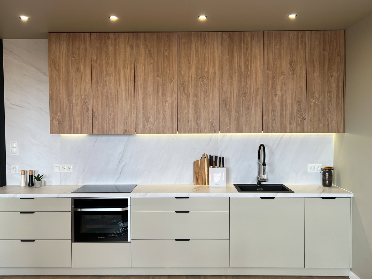 Nowoczesna kuchnia z drewnianymi szafkami wiszącymi, marmurowym backsplashiem, beżowymi szafkami dolnymi z czarnymi uchwytami, czarnym zlewem i indukcyjną płytą grzewczą.