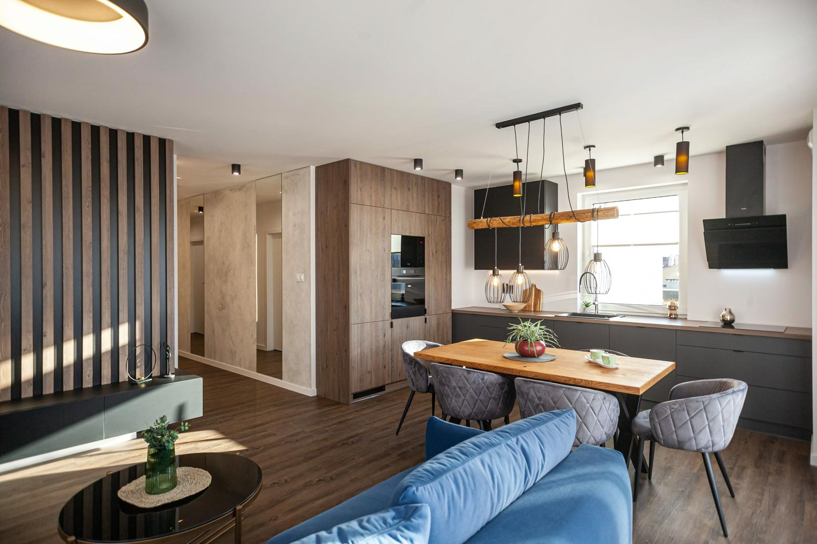 Nowoczesne wnętrze z drewnianą podłogą, ścianą z pionowych paneli, lustrzaną ścianą, szarą zabudową kuchenną, drewnianym stołem i niebieską sofą.