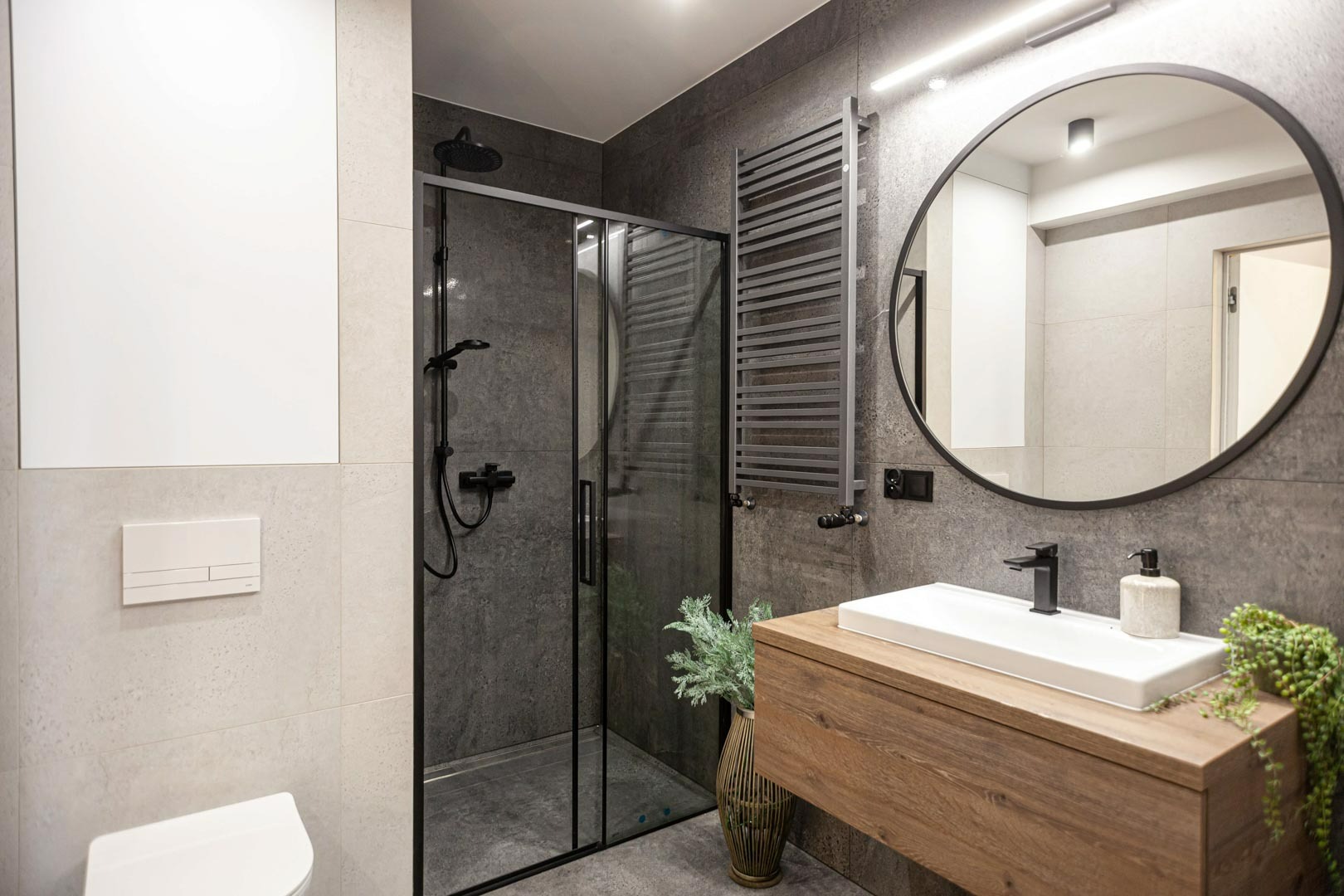 Nowoczesna łazienka z kabiną prysznicową typu walk-in z czarnymi profilami, okrągłym lustrem w czarnej ramie, drewnianą szafką pod umywalką i szarymi płytkami.