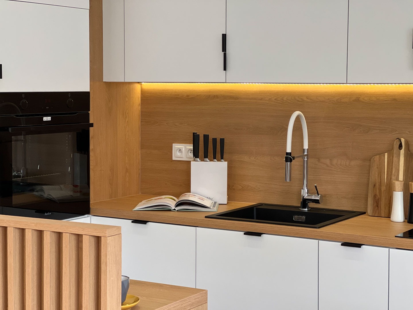 Nowoczesna kuchnia z białymi szafkami, drewnianym blatem i oświetleniem LED pod szafkami wiszącymi, czarny zlew i bateria z elastyczną wylewką, na blacie otwarta książka kucharska i blok z nożami.