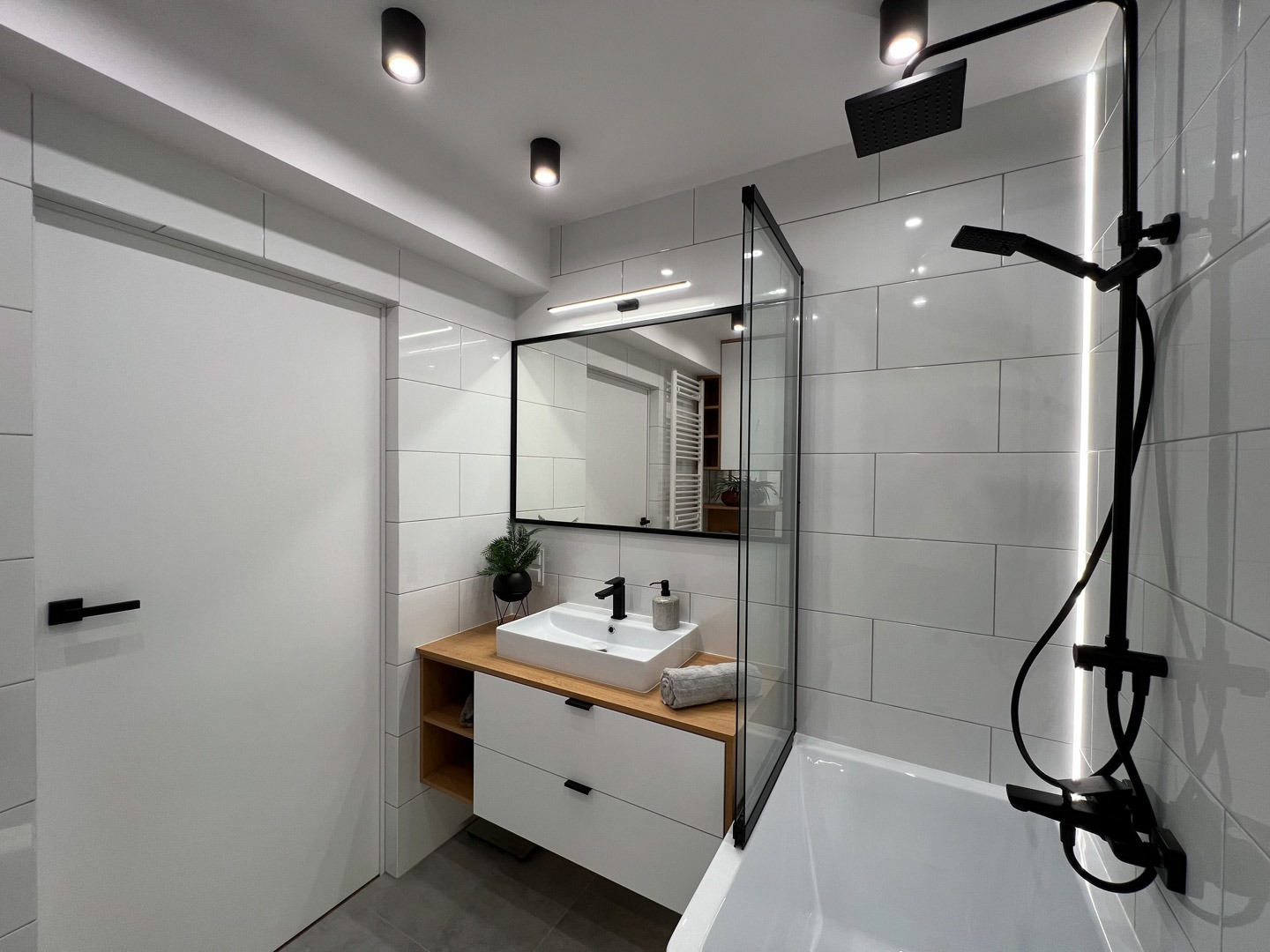 Nowoczesna łazienka z białymi płytkami, czarną armaturą i drewnianym blatem pod umywalką, oświetlenie punktowe sufitowe i listwa LED za kabiną prysznicową.