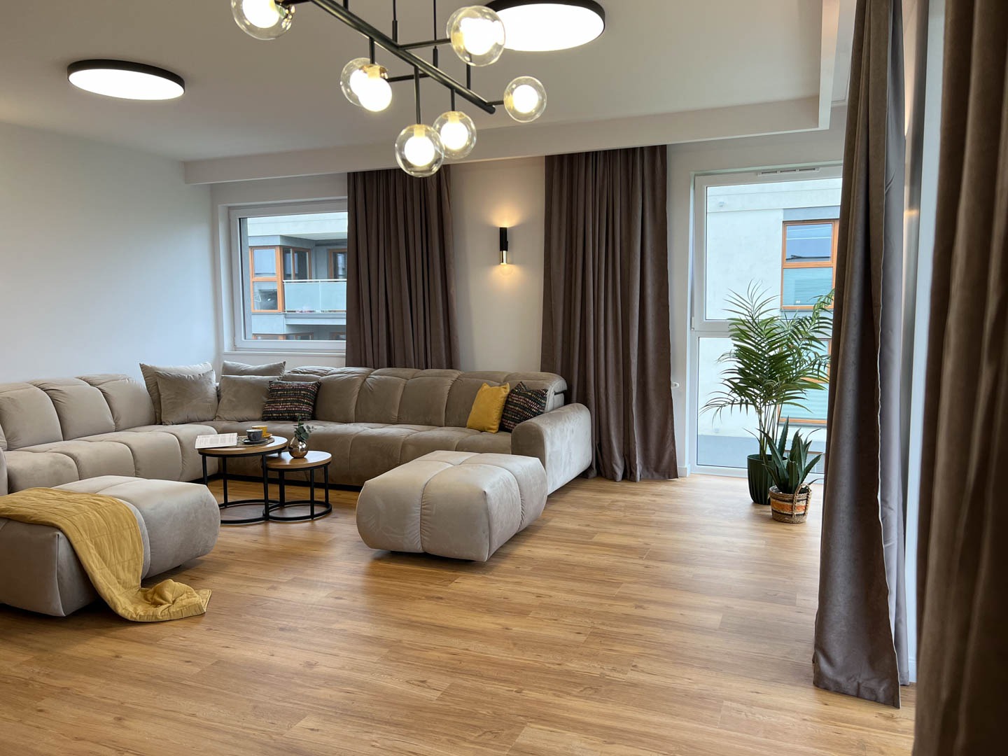 Jasny salon z narożną sofą w odcieniach beżu, drewnianą podłogą i nowoczesnym oświetleniem sufitowym z kulistymi kloszami, widok na okno z rośliną doniczkową i zasłonami.