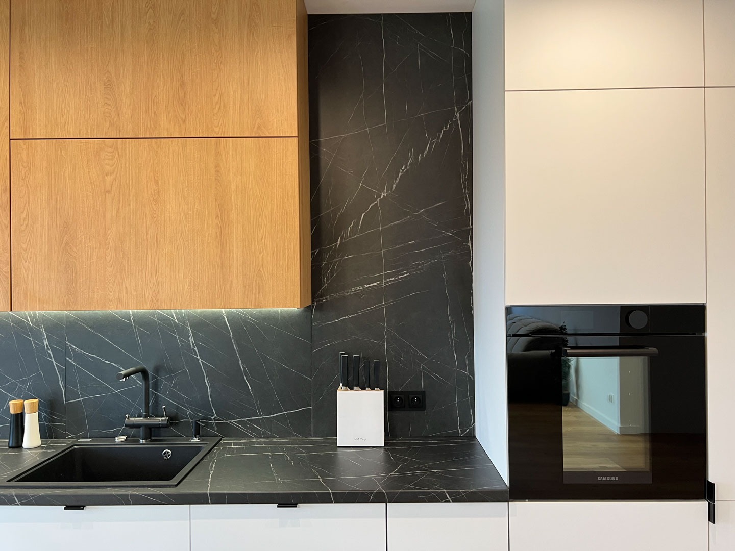 Nowoczesna kuchnia z drewnianymi i białymi szafkami, czarnym blatem z marmurowym wzorem i wbudowanym piekarnikiem Samsung, oświetlona listwą LED pod szafkami wiszącymi.