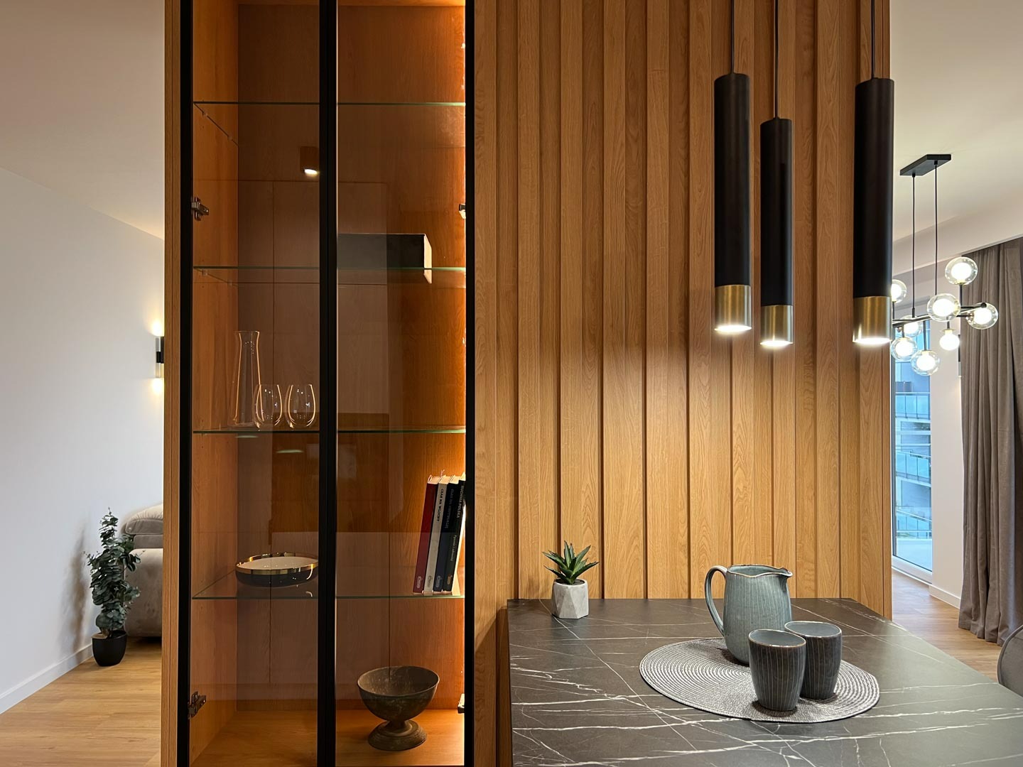 Nowoczesne wnętrze z drewnianą ścianą z pionowych paneli, oświetleniem punktowym i przeszkloną witryną z dekoracyjnymi przedmiotami, widok na salon z sofą.