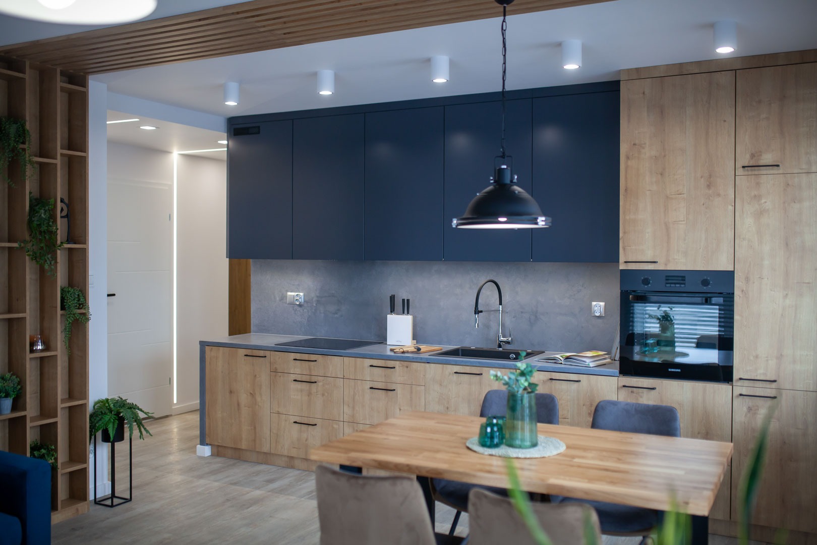 Nowoczesna kuchnia z drewnianymi szafkami, ciemnoniebieskimi górnymi szafkami, szarą ścianą nad blatem, czarnym zlewem i lampą wiszącą, z drewnianym stołem jadalnym i krzesłami w pierwszym planie.
