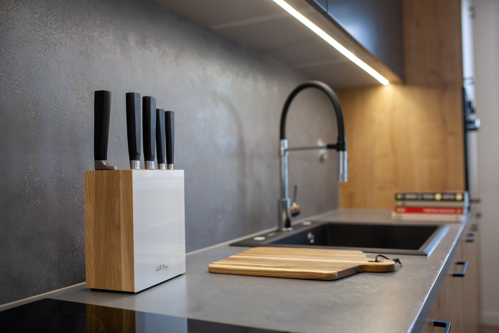 Nowoczesna kuchnia z szarym blatem, czarnym zlewem, drewnianą deską do krojenia i zestawem noży w bloku, oświetlona listwą LED pod szafkami.