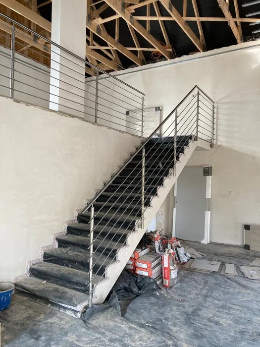 Wewnętrzne schody z czarnymi stopniami i balustradą ze stali nierdzewnej w trakcie montażu, widoczne materiały budowlane i surowy stan wykończenia wnętrza.