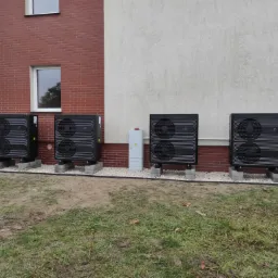 Cztery czarne pompy ciepła zamontowane na betonowych podstawach przed budynkiem z ceglaną i jasną elewacją, widoczna skrzynka elektryczna i trawnik z elementami ziemi.