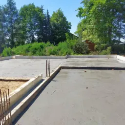 Wylany betonowy fundament pod budynek, z wystającymi prętami zbrojeniowymi, widok z g&oacute;ry na działkę budowlaną z zielenią w tle.