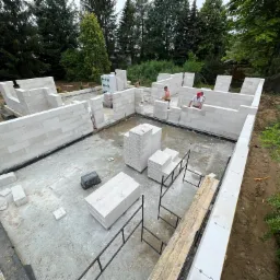 Budowa domu jednorodzinnego: wznoszenie ścian z białych bloczków, widoczni dwaj pracownicy w trakcie murowania, palety z materiałem budowlanym na fundamencie, zieleń w tle.