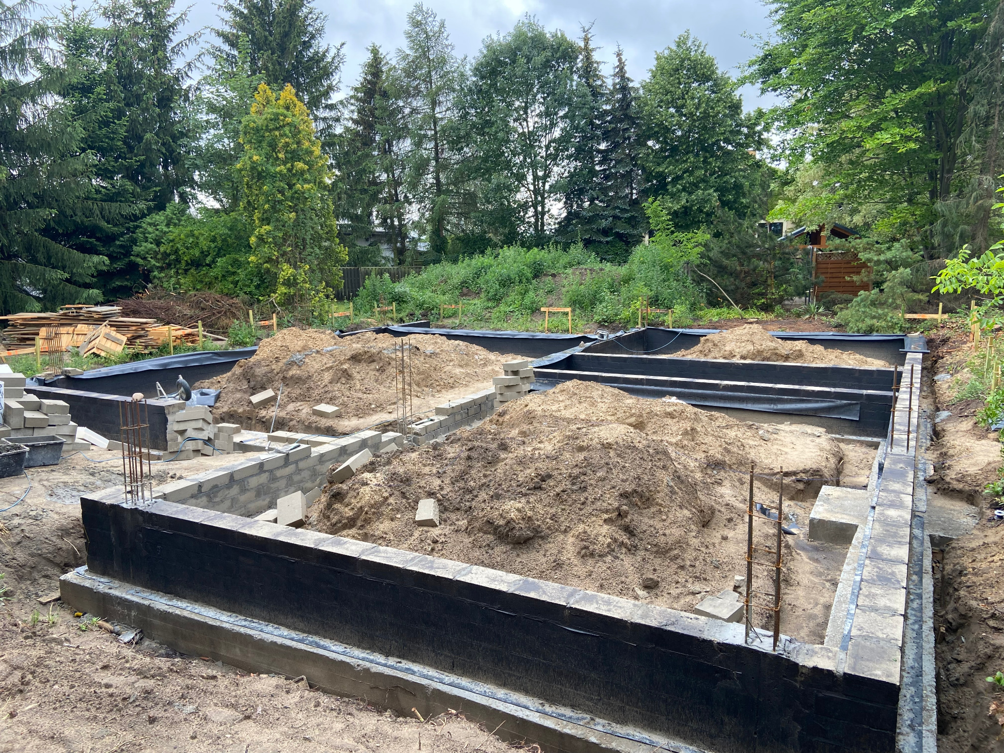 Etap budowy fundamentów: zalane betonem ściany z bloczków, zabezpieczone czarną folią, z widocznymi sterczącymi prętami zbrojeniowymi i stertami ziemi wewnątrz obrysu, w tle drzewa.