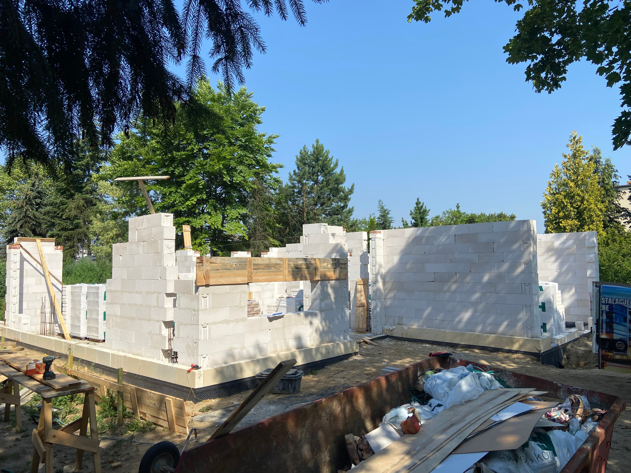 Wznoszenie ścian domu z bloczków betonu komórkowego, widoczny fragment zbrojenia, drewniane stemple i szalunki, w tle zieleń drzew i niebieskie niebo.