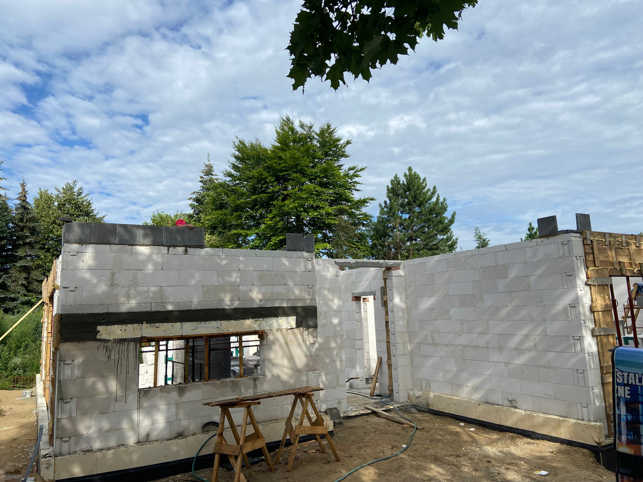 Budowa domu z białych bloczków, widoczne ściany w trakcie murowania, drewniane kozły i częściowo zamontowane okno, tło z drzewami i błękitnym niebem z chmurami.