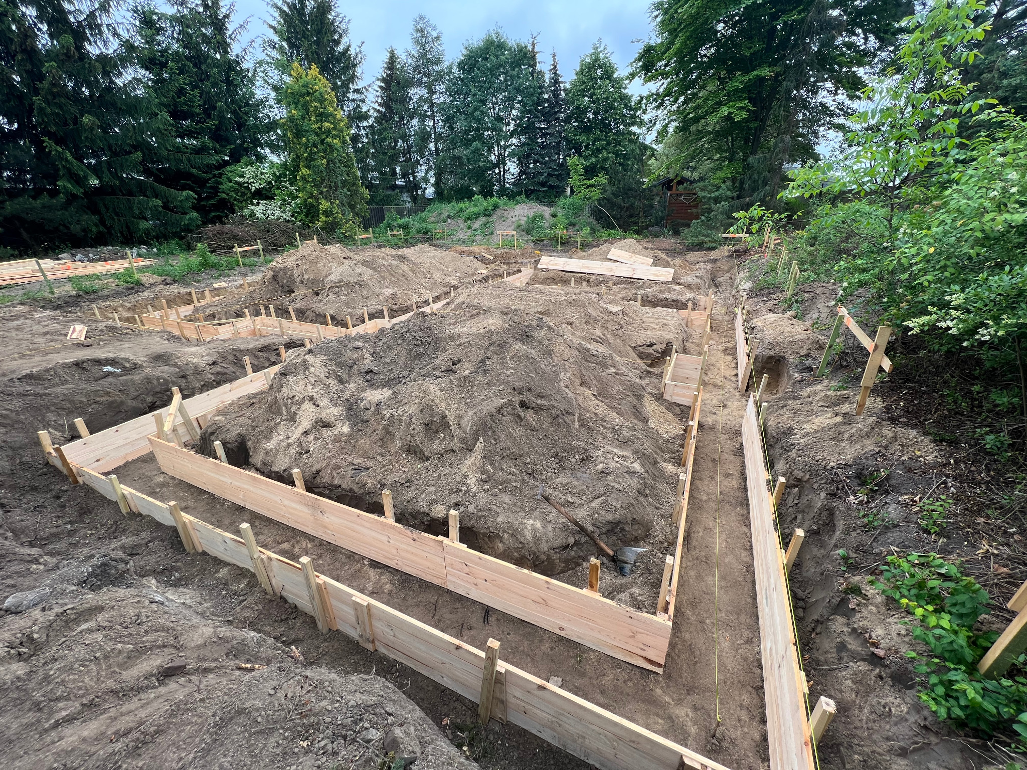Wykonane deskowanie pod fundamenty domu jednorodzinnego na działce z zielenią, widoczne wykopy i pryzmy ziemi.