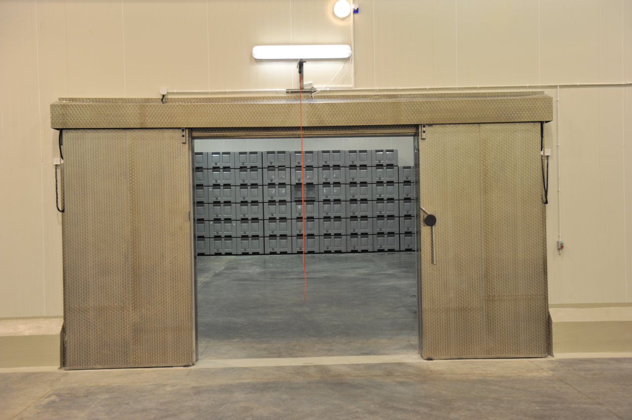 Otwarta, przesuwna brama do chłodni z widocznymi pojemnikami wewnątrz, oświetlona lampą jarzeniową.