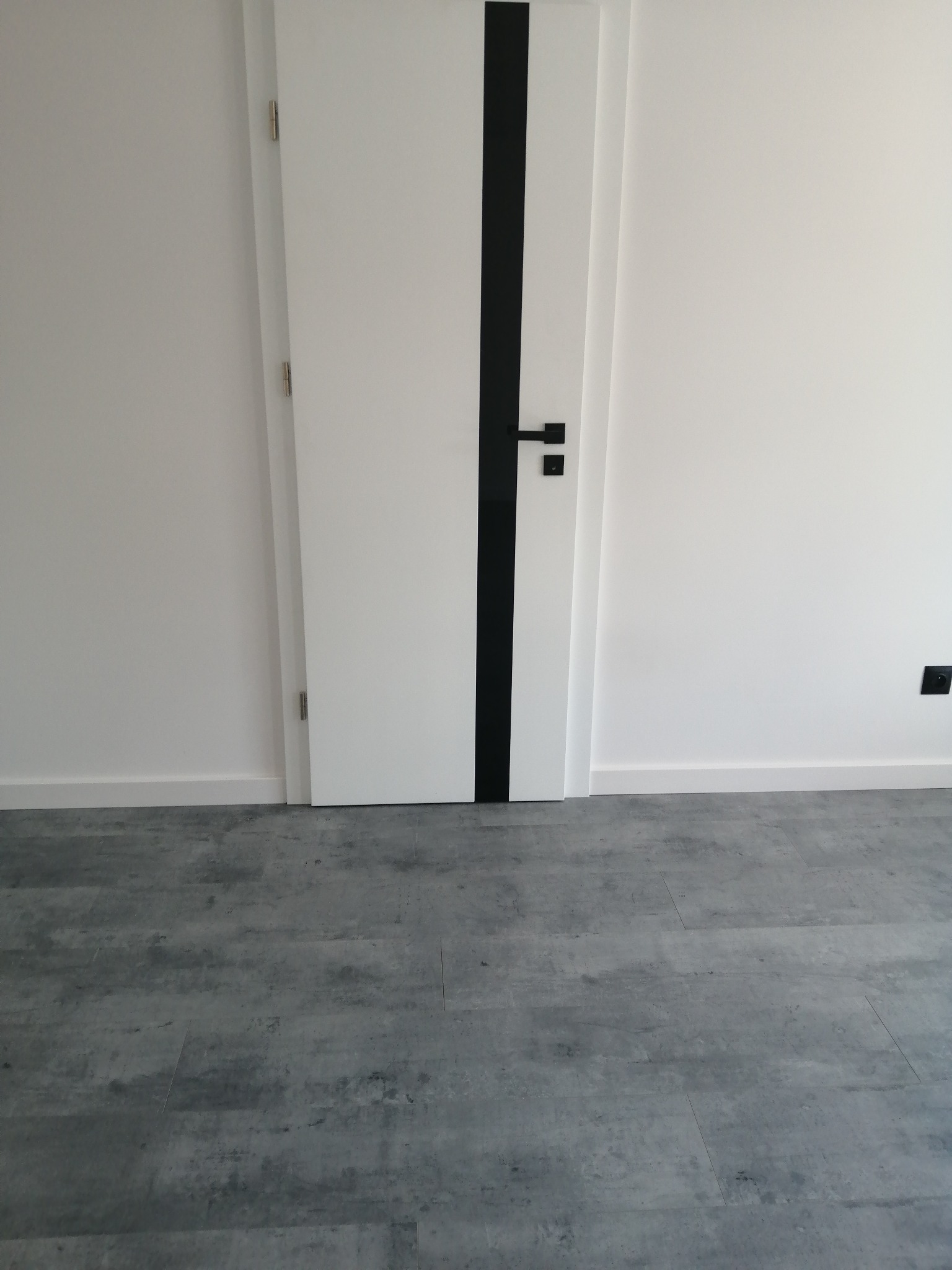 Szara podłoga z paneli imitujących beton, biała ściana z przypodłogową listwą i białe drzwi z czarnym pionowym paskiem dekoracyjnym i czarną klamką.