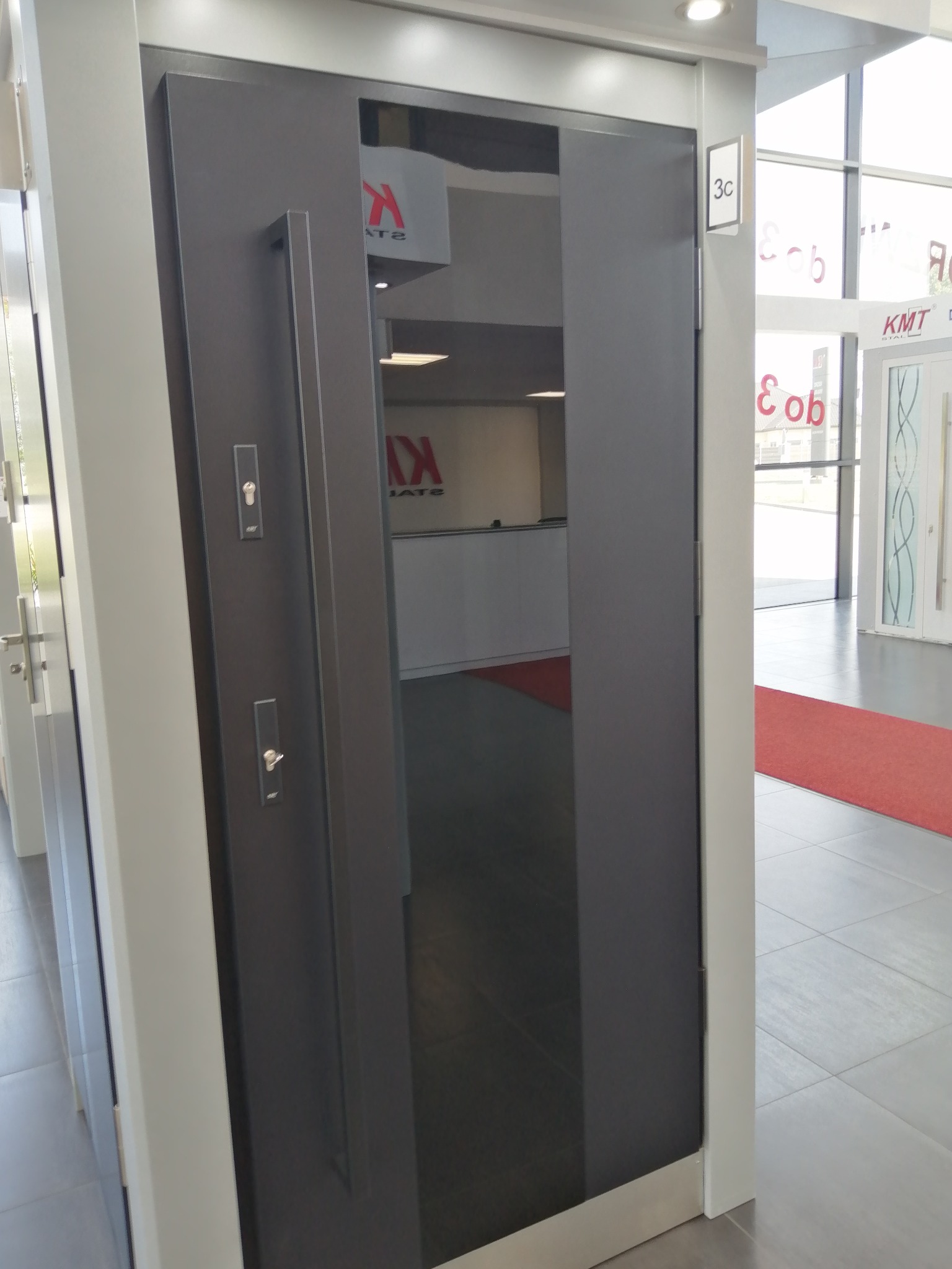 Nowoczesne, grafitowe drzwi wejściowe z długim, pionowym uchwytem i dwoma zamkami, widoczne w salonie ekspozycyjnym z czerwonym dywanem i innymi modelami drzwi w tle.