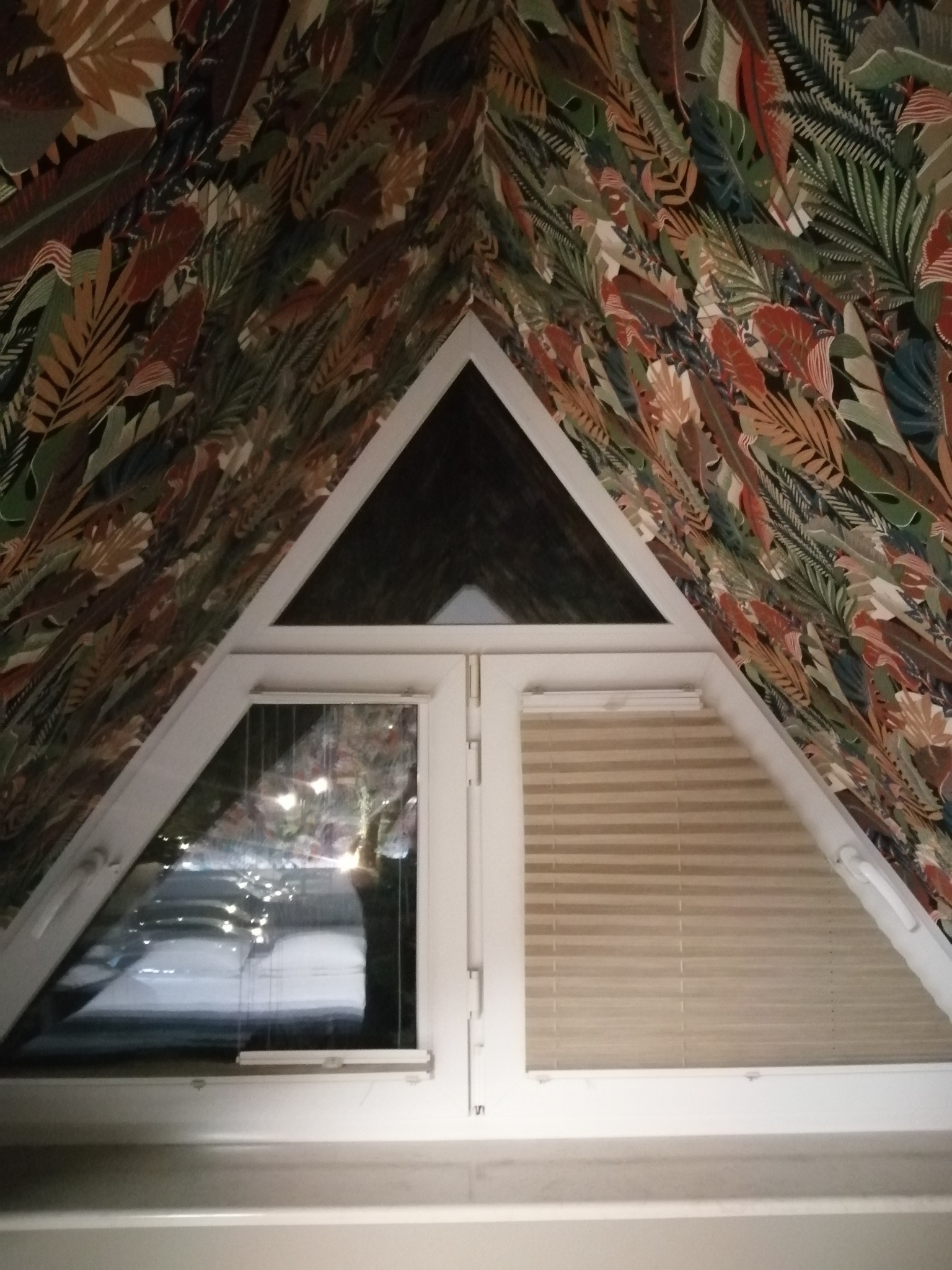 Trójkątne okno PCV na poddaszu z tapetą w motywy liści, roleta pozioma w jednym oknie, widok na zewnątrz przez drugie okno.