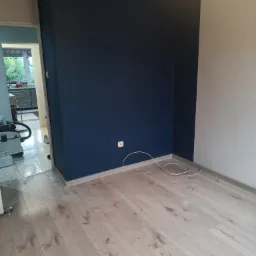 Pomalowany róg pokoju: ściana w kolorze głębokiego granatu sąsiaduje z białą ścianą, jasna podłoga z paneli, widoczny kabel wystający ze ściany przy podłodze, w tle widok na kuchnię.