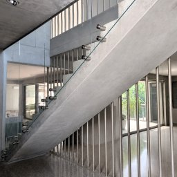 Vitrada sp. z o.o. - Nowoczesne schody z surowego betonu z przezroczystą osłoną mocowaną do boku na metalowych dystansach. W tle widoczne duże okna i zieleń.