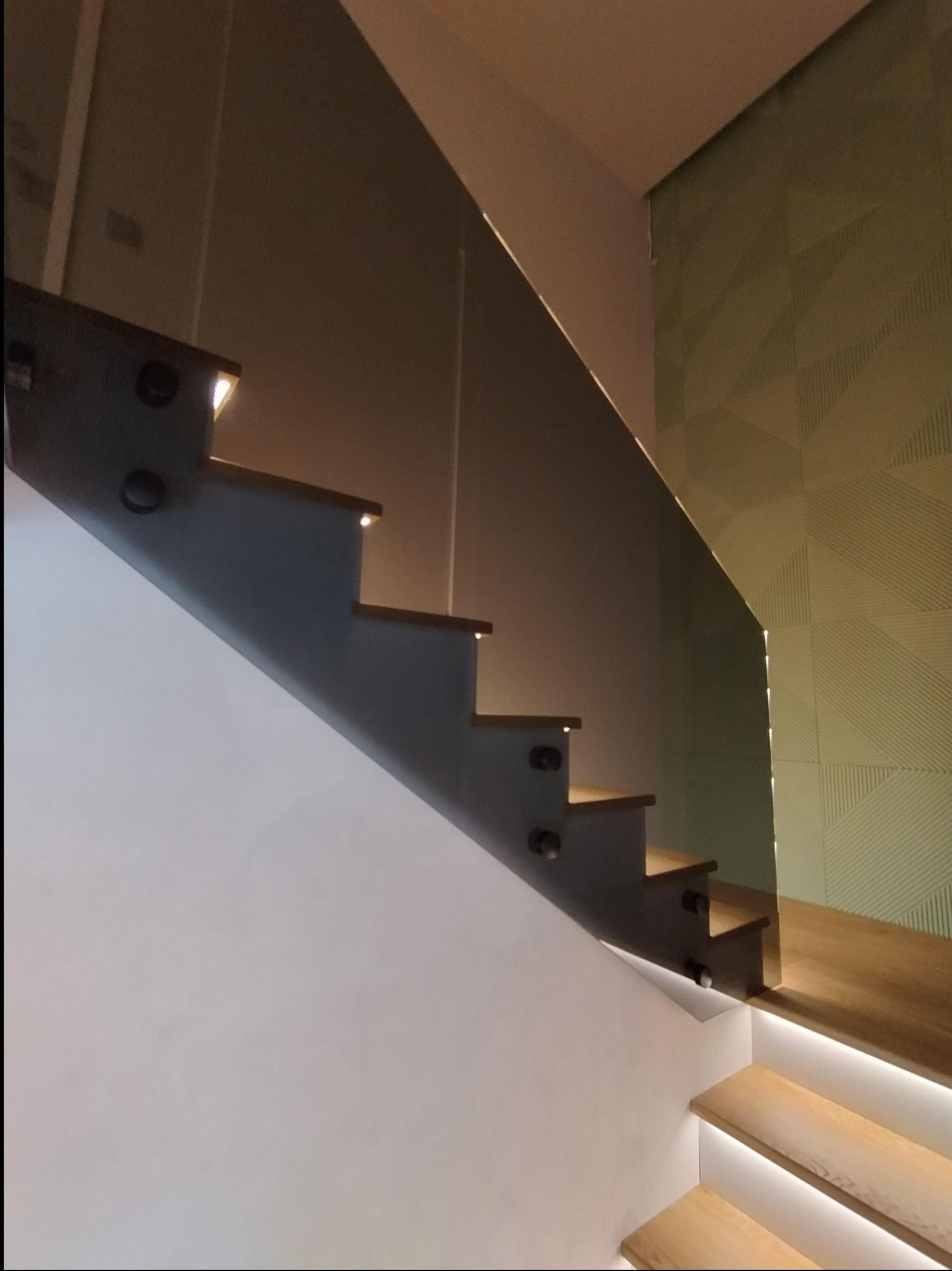 Szklana barierka zabezpieczająca schody z drewnianymi stopniami, oświetlonymi od spodu, zamocowana za pomocą punktowych uchwytów, na tle ściany z geometrycznym wzorem.