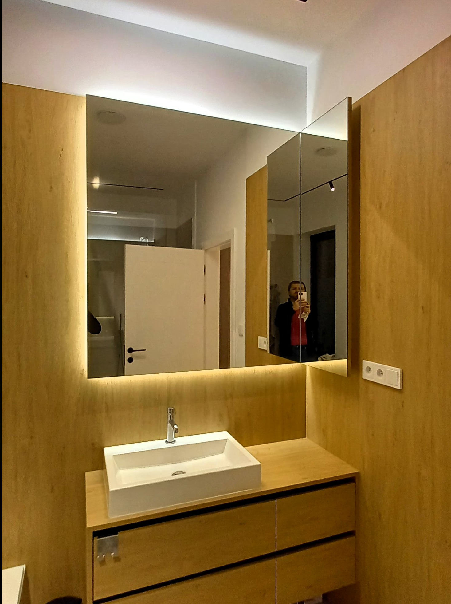 Łazienka w odcieniach drewna z podświetlanym lustrem LED, odbicie osoby robiącej zdjęcie w lustrze szafki.