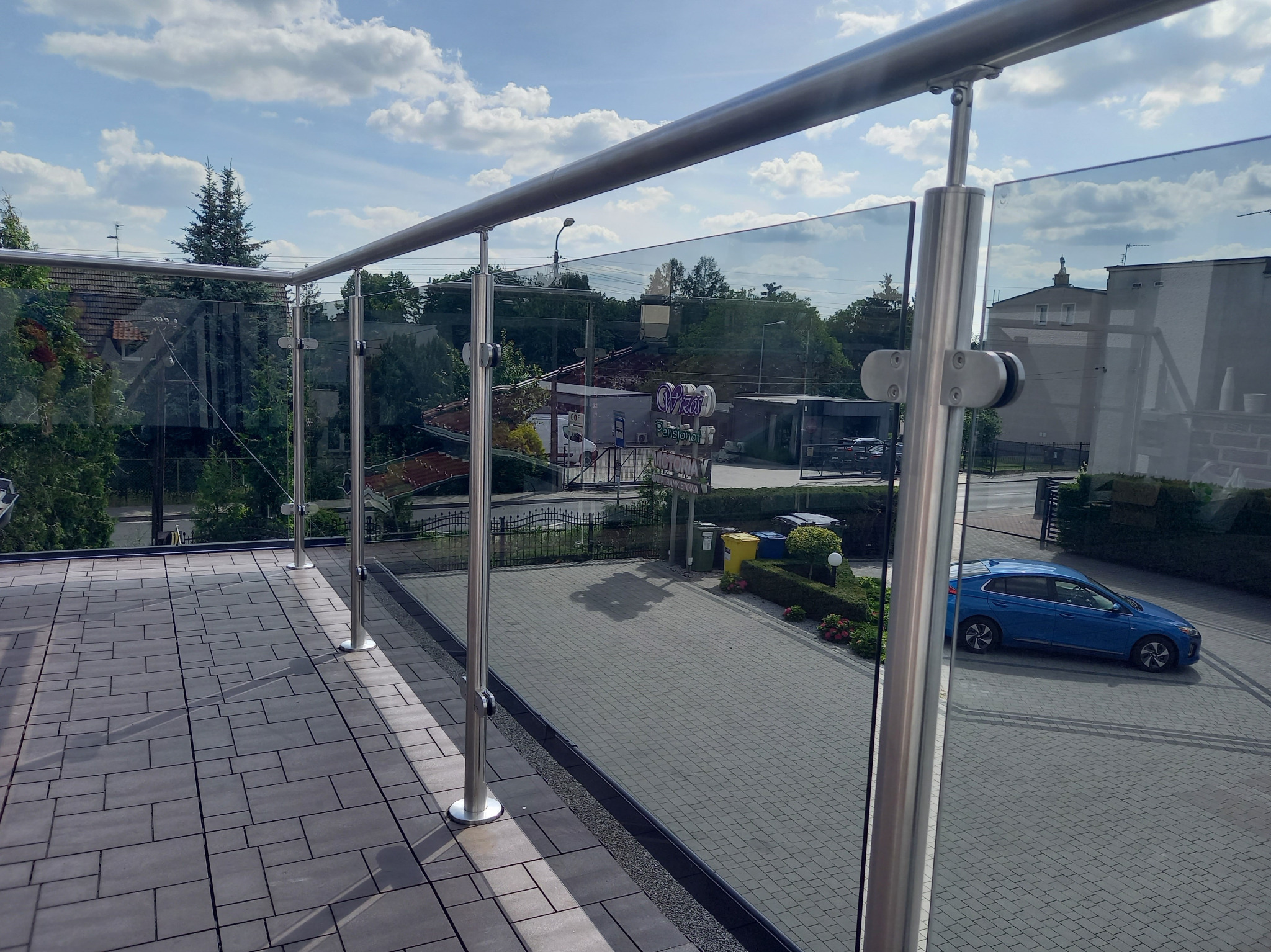 Szklana balustrada balkonowa z metalowymi słupkami, widok z góry na szare płytki tarasowe i niebieski samochód odbijający się w szkle.