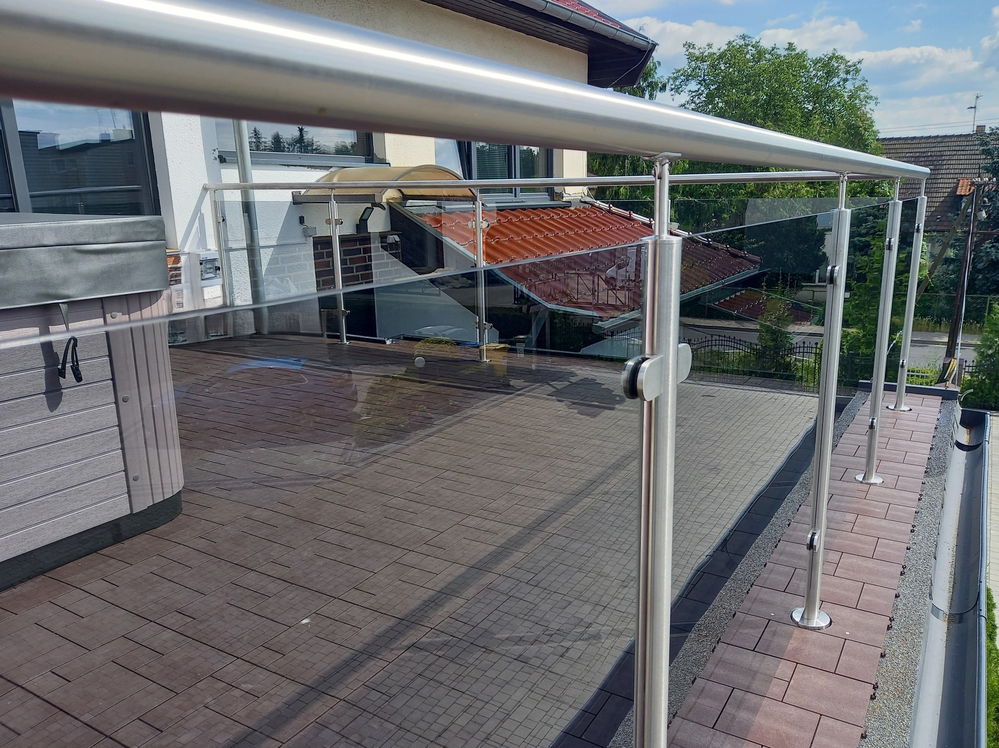 Nowoczesna balustrada ze stali nierdzewnej i szkła na tarasie z drewnopodobną podłogą, widok z góry na dom z czerwoną dachówką w tle.