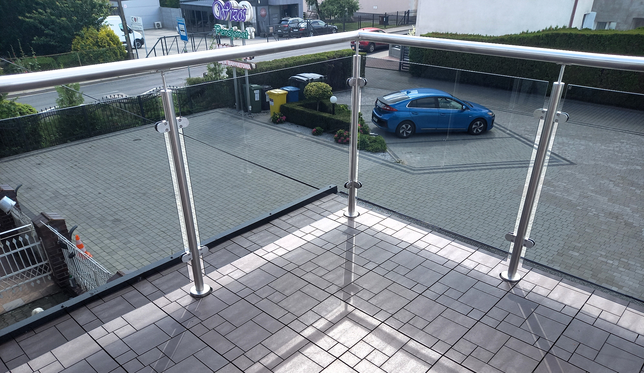 Szklana balustrada z metalowymi słupkami na tarasie z szarej kostki, widok z góry na podjazd z zaparkowanym niebieskim samochodem.