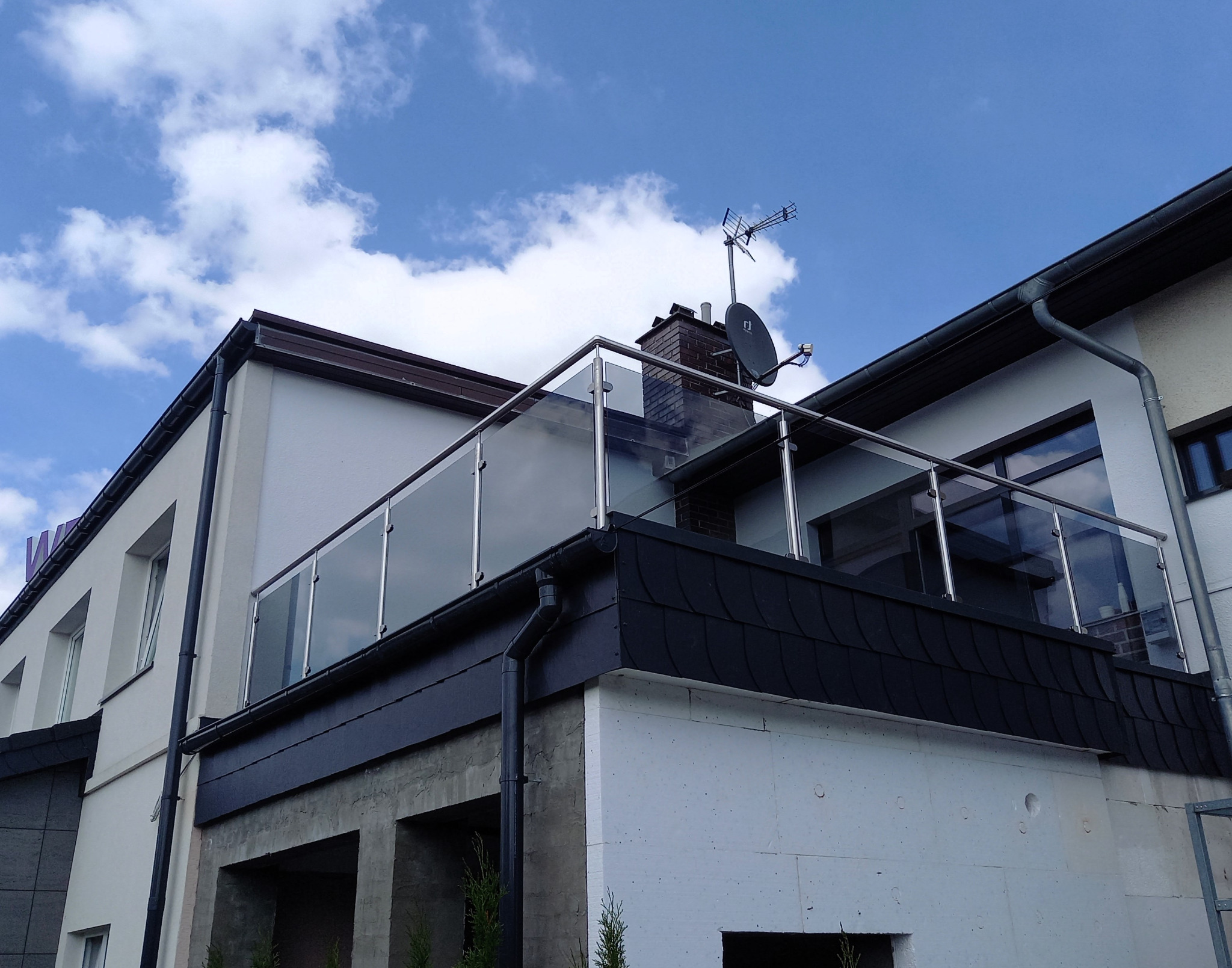 Zewnętrzna balustrada ze szkła hartowanego i stali nierdzewnej na balkonie budynku, widoczna antena satelitarna na dachu.