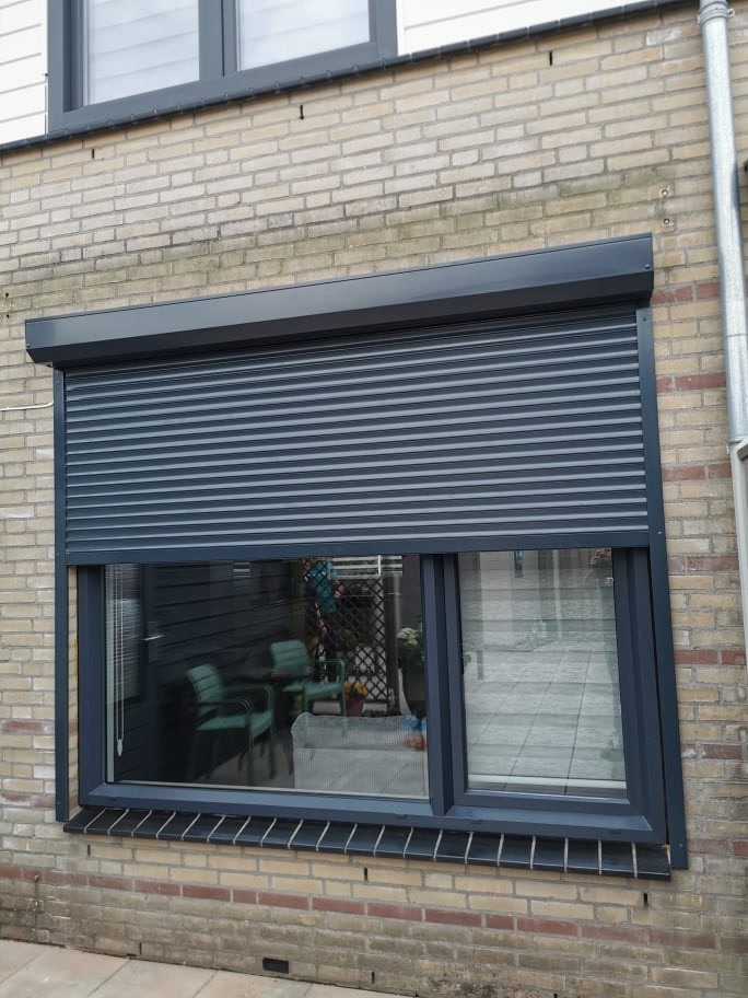 Szare okno z roletą zewnętrzną zamontowaną na ceglanej elewacji budynku. Widoczne odbicie wnętrza pokoju w szybie.