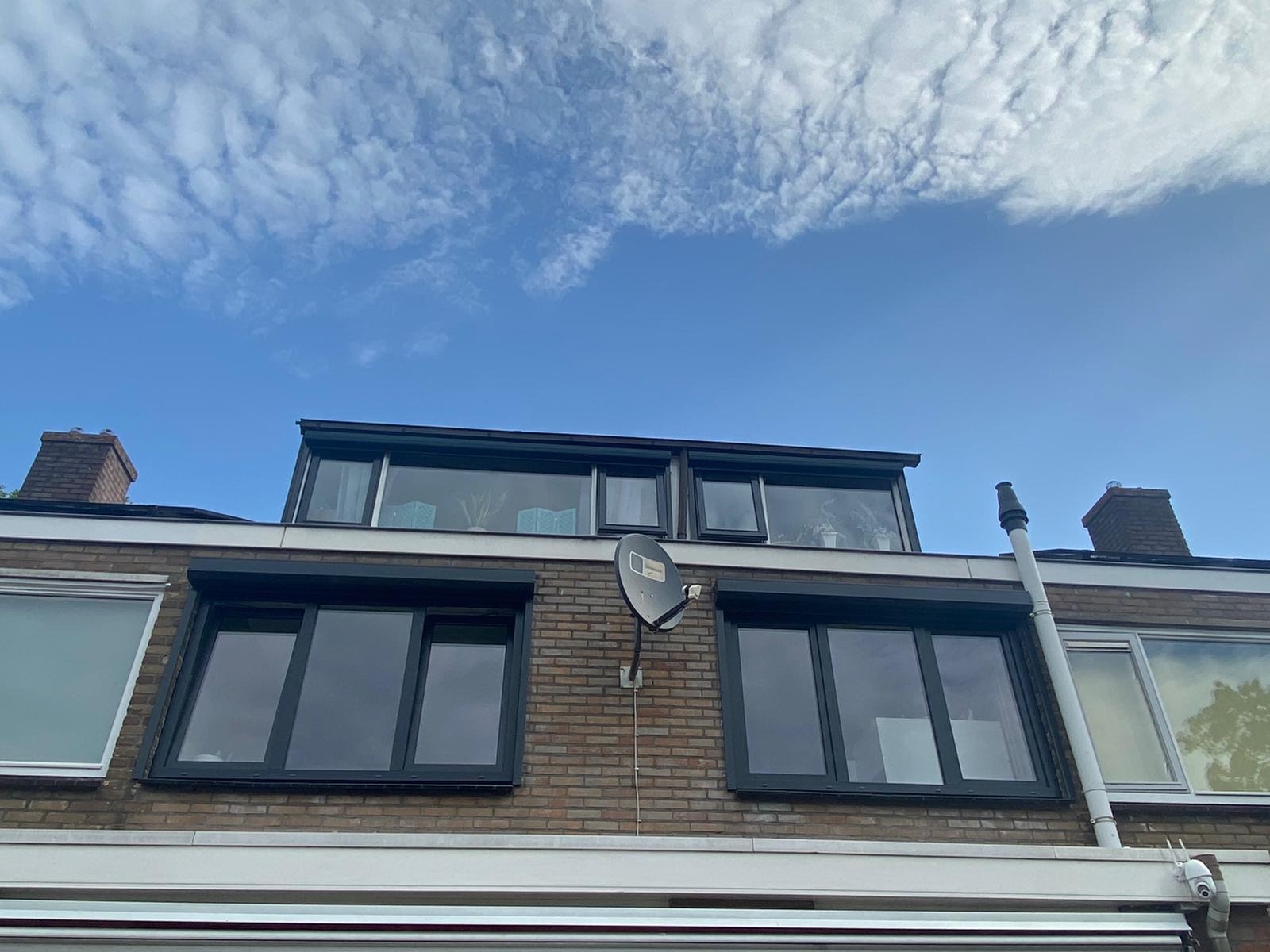 Elewacja domu z cegły z oknami z czarnymi ramami i opuszczonymi roletami zewnętrznymi, na ścianie antena satelitarna, w tle błękitne niebo z chmurami.