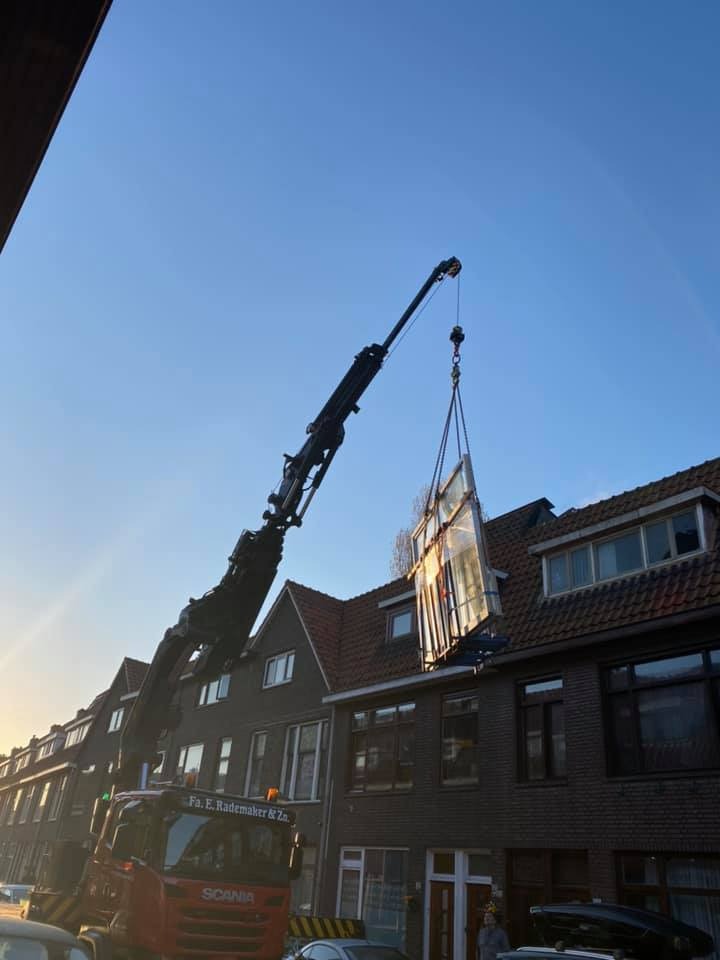 Montaż okna dźwigiem na tle kamienic z czerwonej cegły w słoneczny dzień.