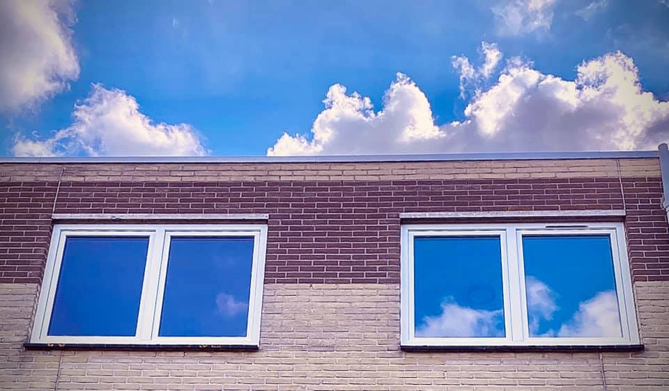 Dwa białe okna na ceglanej ścianie budynku, z odbijającym się błękitnym niebem z chmurami w szybach.