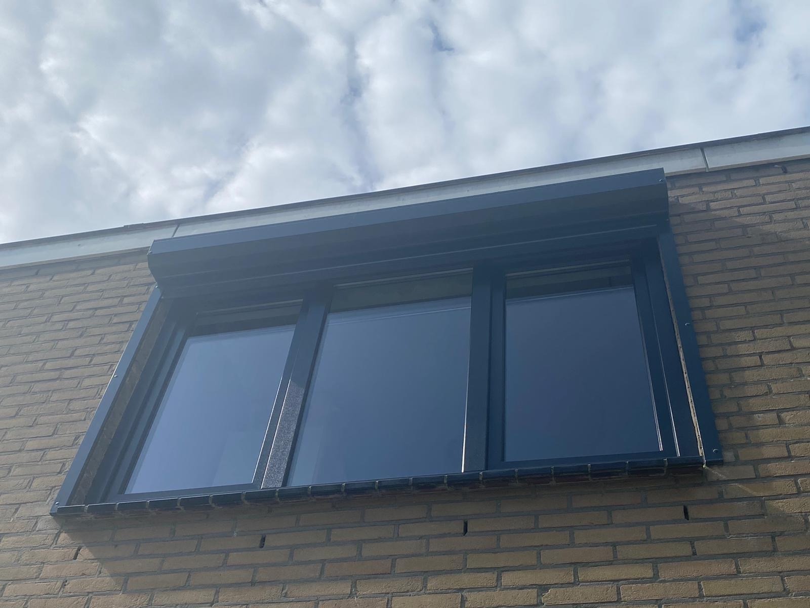 Trzyczęściowe okno z ciemną ramą i zwiniętą roletą zewnętrzną na ceglanej ścianie budynku, widok z dołu w pochmurny dzień.
